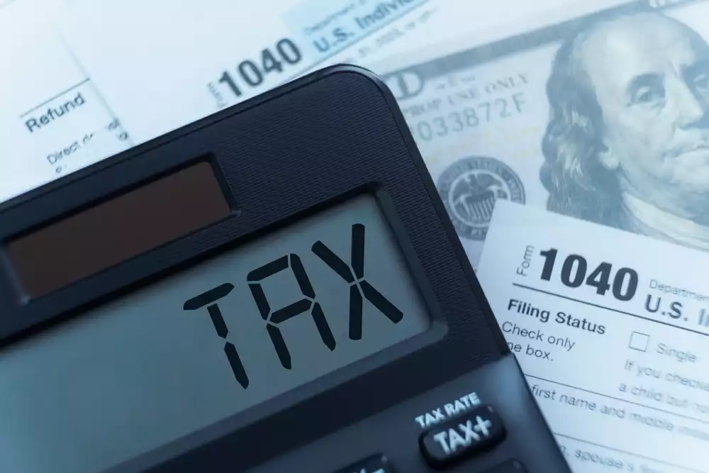 The word “Tax” spelled out on a calculator next to an IRS 1040 form and a 100 dollar bill