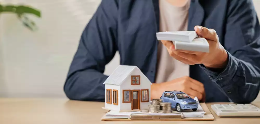 Man holds pile of money over a model of a house and car representing the concept of a property tax payment plan
