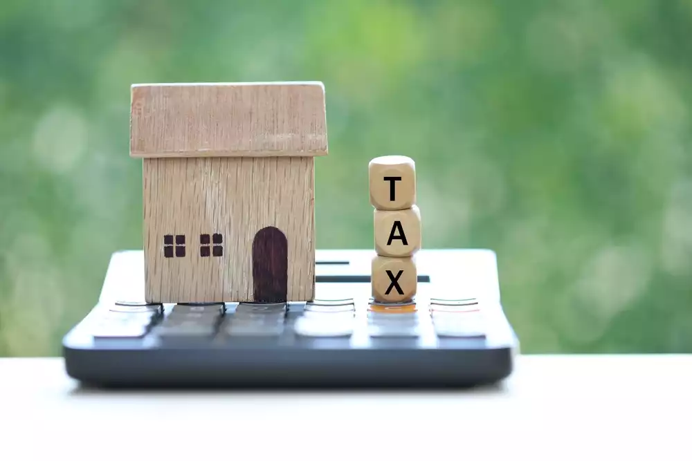 “Tax” spelled out on dice next to a small model of a wooden home placed on top of a calculator