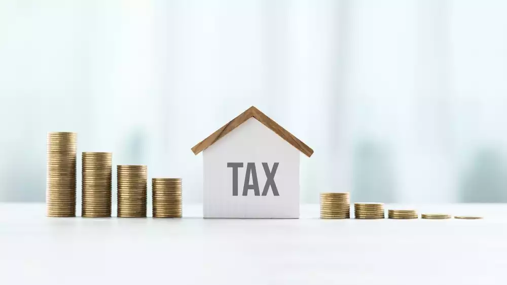 Model sized home with the word “tax” written on it in the middle of a line of coin stacks that are slowly decreasing in height representing the concept of help paying property taxes in TX